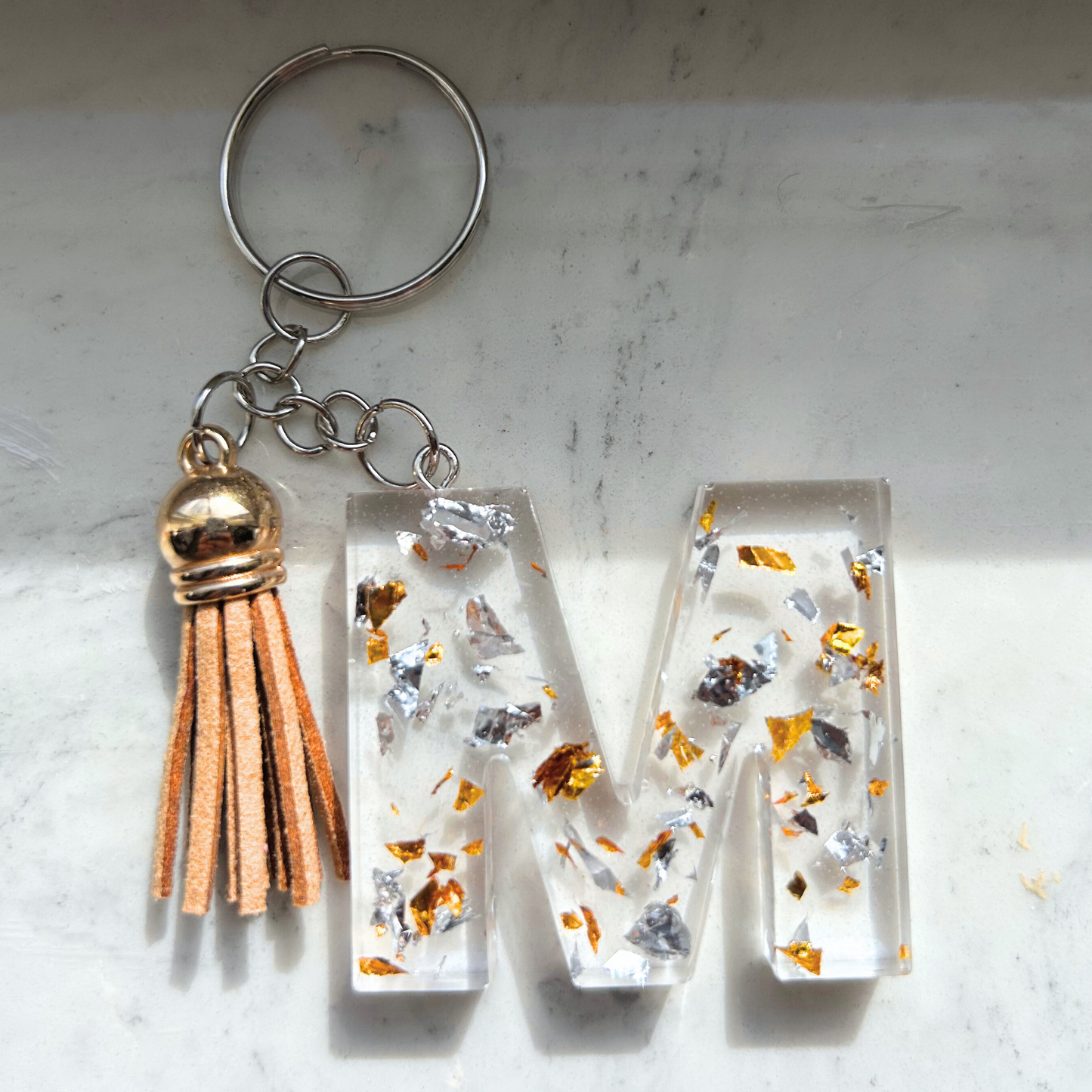 Custom resin keychain with the letter "M", transparent with golden and silver flakes and a color assorted mini tassel.