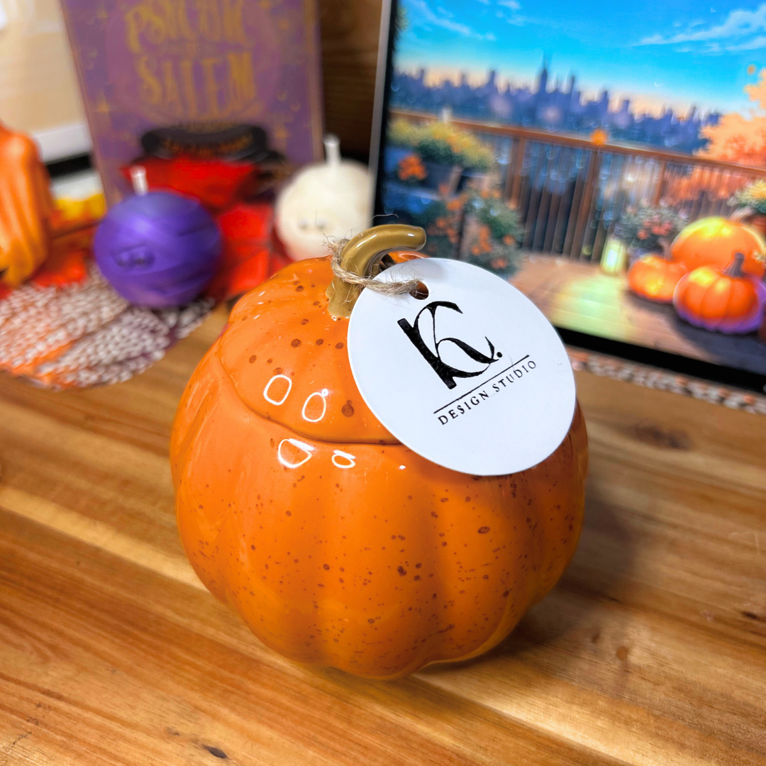 Pumpkin-shaped candle with a tag on a wooden surface, with Halloween-themed decorations in the background.