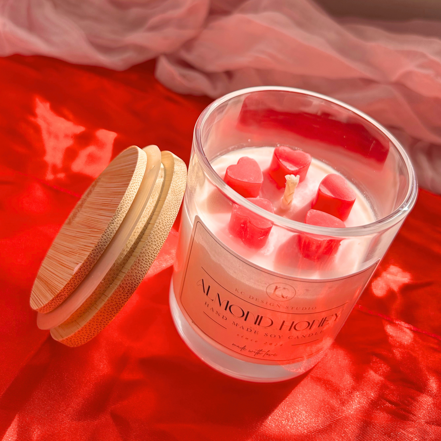 Almond honey candle with pink grapefruit scent, topped with five mini hearts. Lid resting on the side, showcasing a smooth wax finish from a top view.