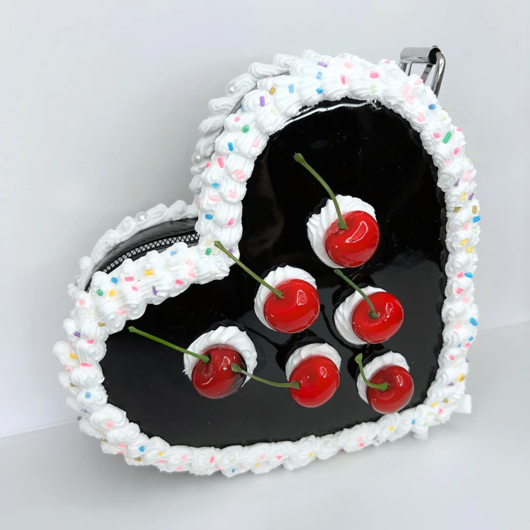 Front view of a black heart-shaped purse with cherry cake design, featuring intricate frosting, sprinkles, and vibrant cherry accents.