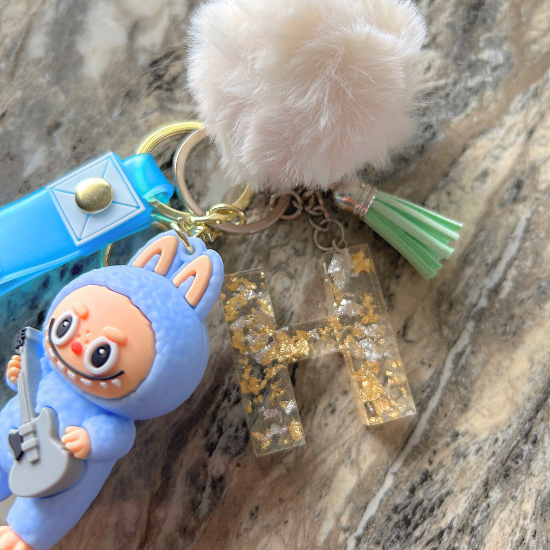 Close-up of custom blue Labubu keychain showing letter charm, gold flakes, pompom, and colorful tassel.