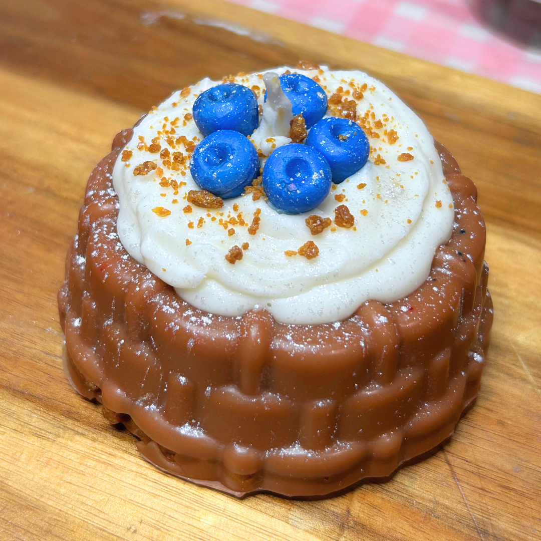 A blueberry shortcake-shaped candle with a brown textured base resembling chocolate cake layers, topped with white frosting and blue berry decorations, sitting on a wooden surface.