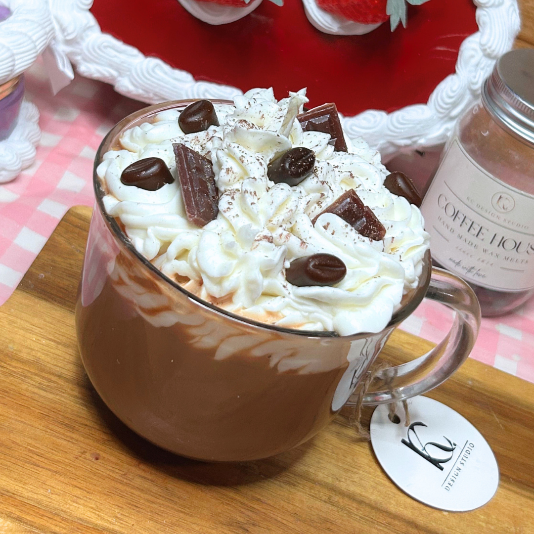 A glass mug candle made with a coffee-style design topped with whipped cream, chocolate chunks, and cocoa powder, set on a wooden board with a branded tag and a candle in the background.