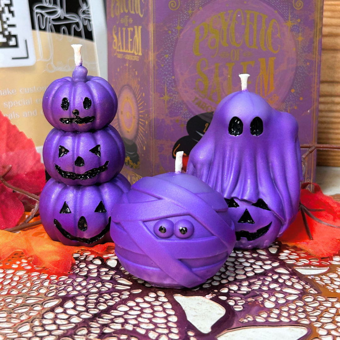Purple Halloween-themed candles shaped like a pumpkin, ghost, and mummy on a decorative tablecloth with a 'Salem' book in the background.