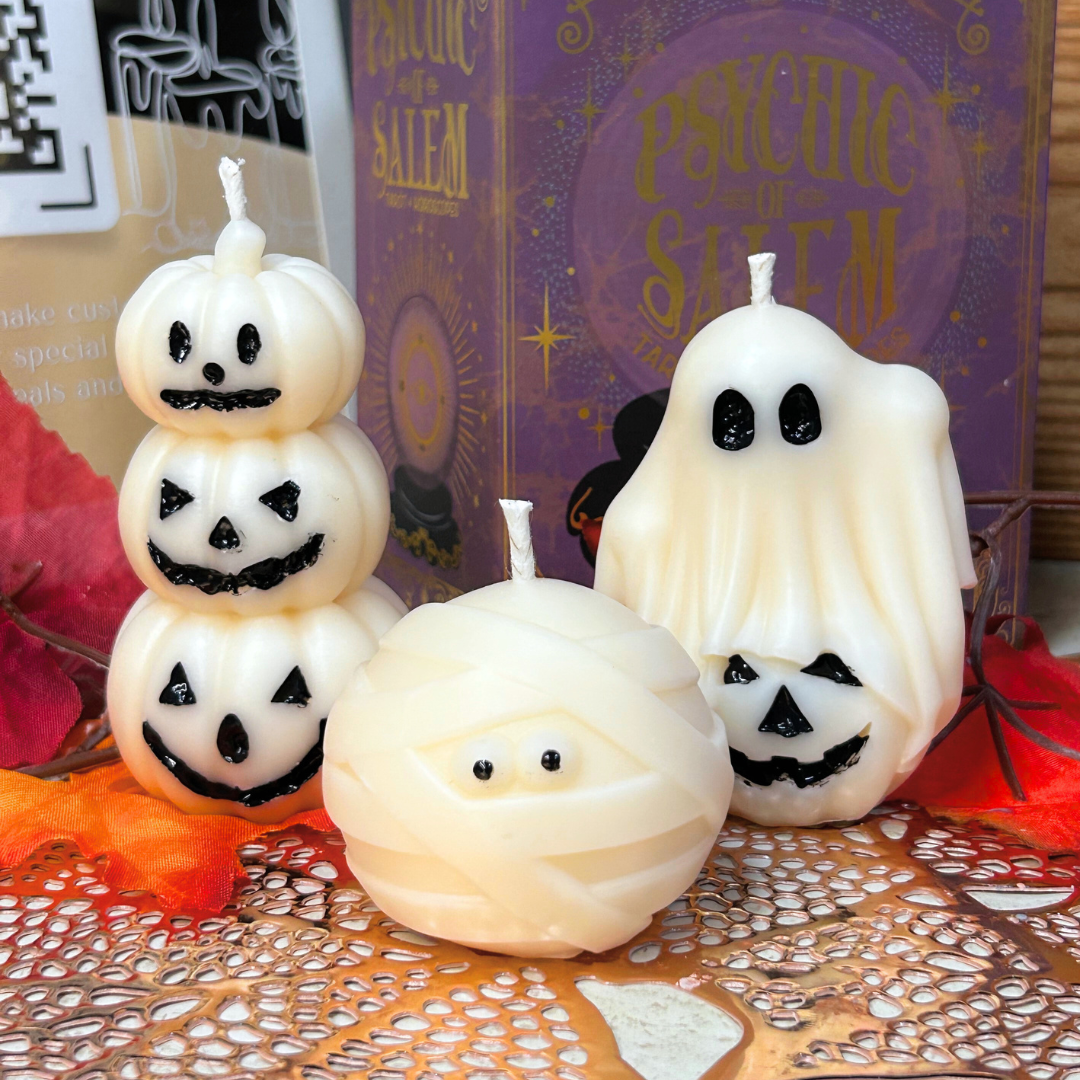 Halloween-themed candles shaped like a ghost, jack-o'-lantern, and mummy on a decorative surface with a mystical book in the background.