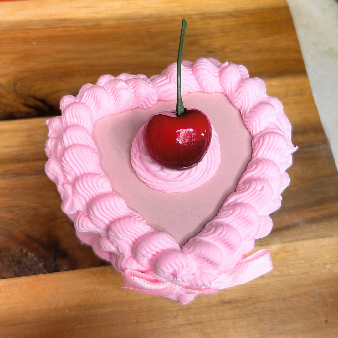 Heart-shaped pink cake trinket box with a cherry on top and pink bow on a wooden surface