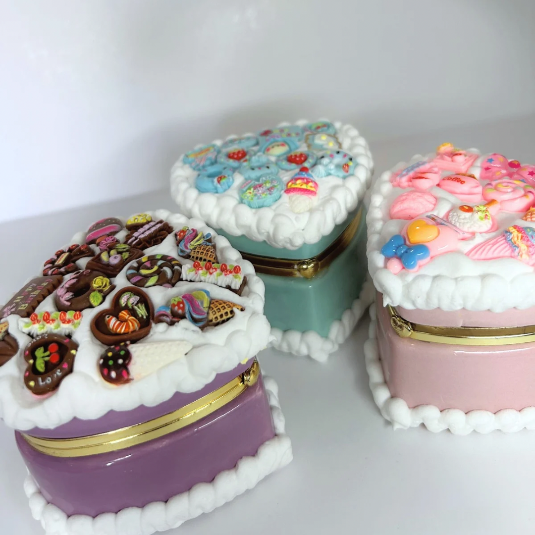 Close-up of three kawaii mini heart-shaped jewelry boxes in pink, blue, and purple, decorated with curvy cream and adorable charms.