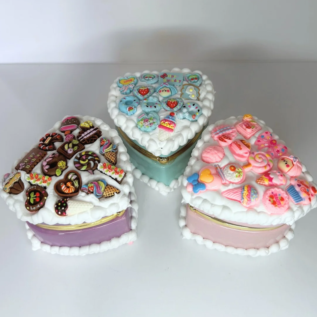 Top view of three kawaii mini heart-shaped jewelry boxes in pink, blue, and purple, decorated with curvy cream and adorable charms.