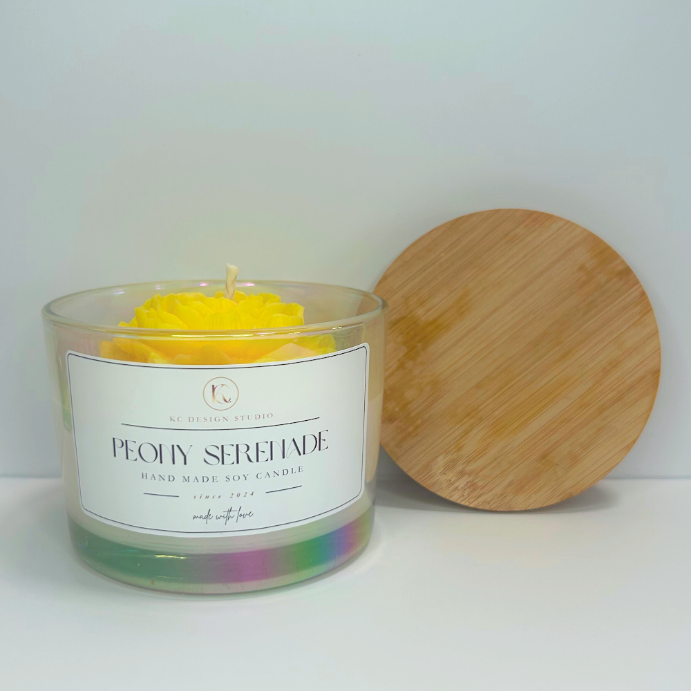 Front view of yellow Peony Serenade Candle with soft floral notes and lid resting on the wall.