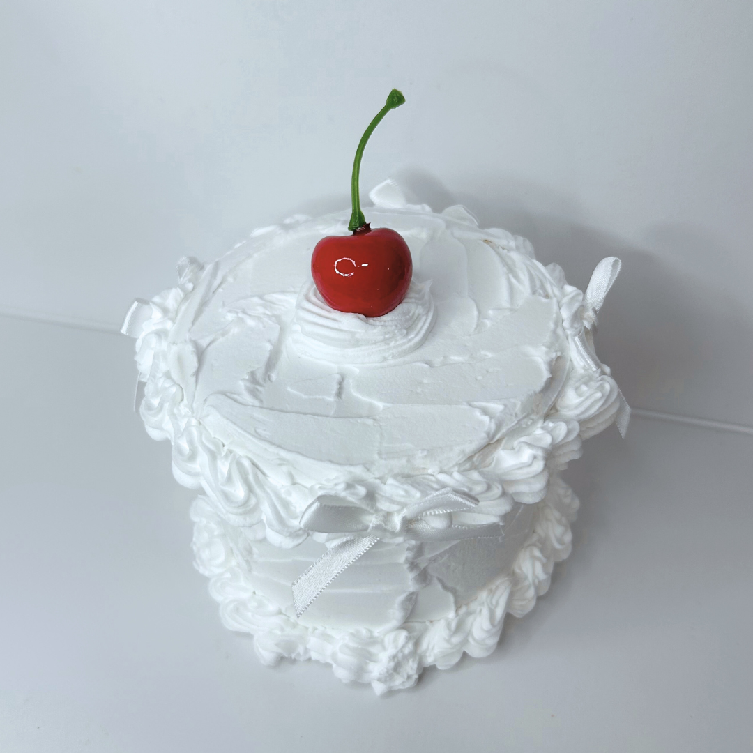 Front view of round cake-themed paper mache box with cherry detail and white bows.