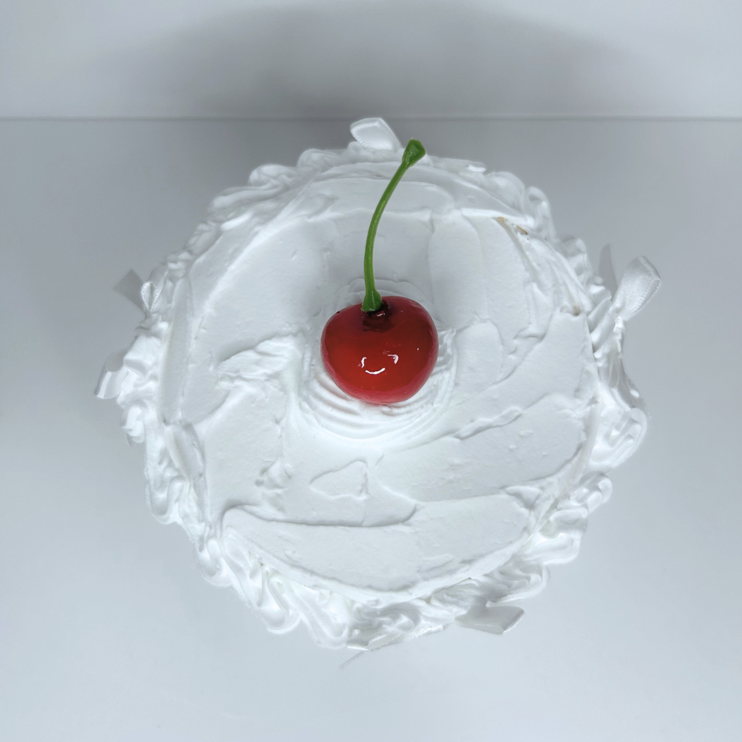 Top view of round cake-inspired storage box with white frosting, cherry center, and four white bows.