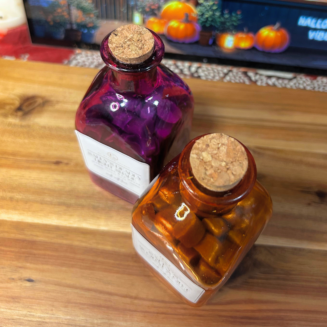 Two wax melt glass bottles with cork lids on a wooden surface, with Halloween-themed decorations in the background.