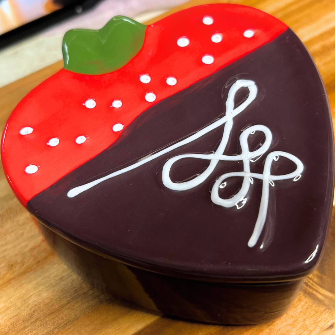 Closed strawberry-shaped trinket box holding chocolate covered strawberry wax melts on a wooden surface with a cozy pink Valentine’s background