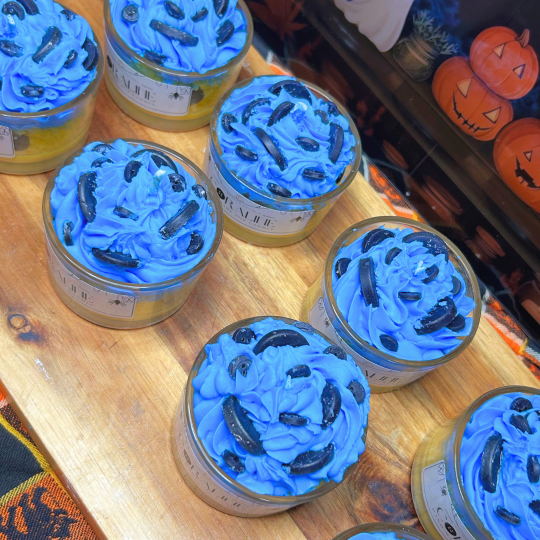 Halloween-themed jar candles with bright blue whipped wax tops and black button decorations, displayed on a wooden board with a Halloween-themed sign in the background.