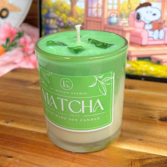Green matcha candle with a label on a wooden surface, with a blurred background of a cartoon character and flowers.
