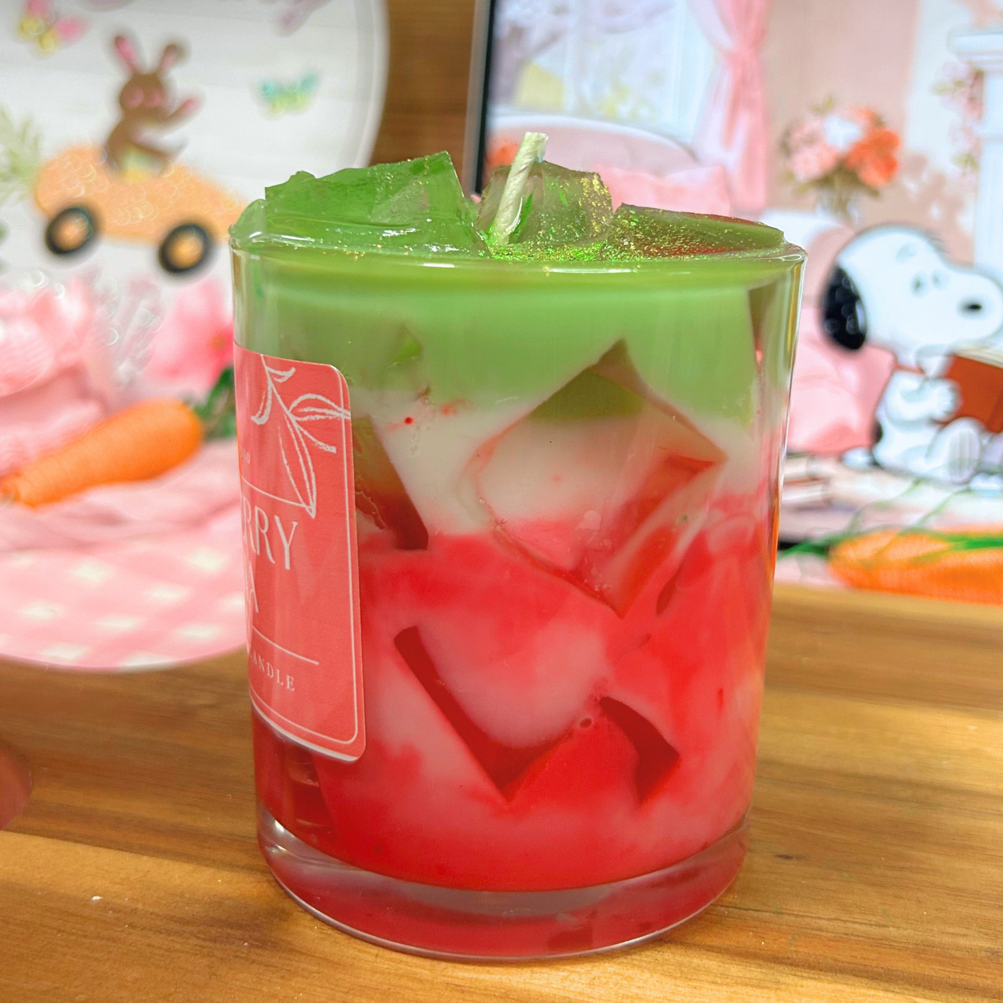 Colorful layered matcha candle in a glass with a brand label, set against a festive background.