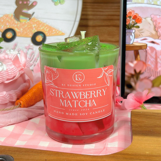 Green candle labeled 'Strawberry Matcha' on a pink checkered surface with blurred background.