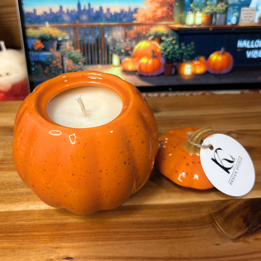 Pumpkin-shaped candle with a lit candle inside on a wooden surface, with a TV displaying Halloween-themed content in the background.