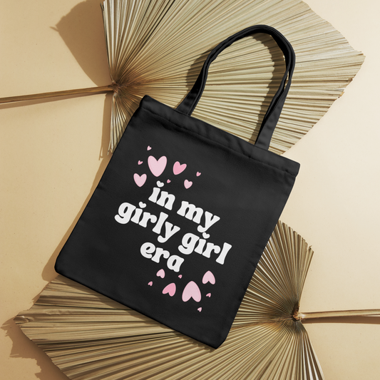 Black canvas tote bag with "In My Girly Girl Era" text and pink floating hearts, perfect for daily use.