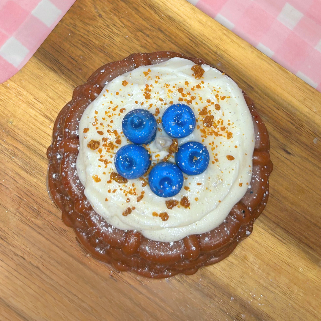 A round, browned blueberry shortcake candle topped with thick white cream, five blueberries, and brown crumb sprinkles, presented on a wooden board with a pink-and-white checkered cloth in the background.