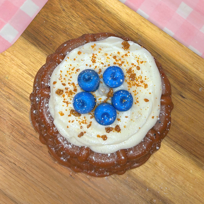 A round, browned blueberry shortcake candle topped with thick white cream, five blueberries, and brown crumb sprinkles, presented on a wooden board with a pink-and-white checkered cloth in the background.