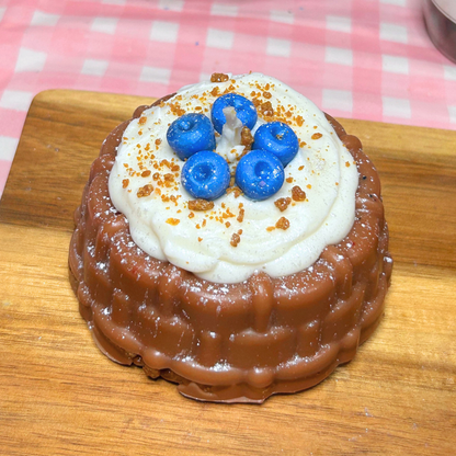A small round shortcake candle on a wooden board, topped with white whipped cream, four blue berries, and brown crumbs, with a pink-and-white checkered cloth in the background.