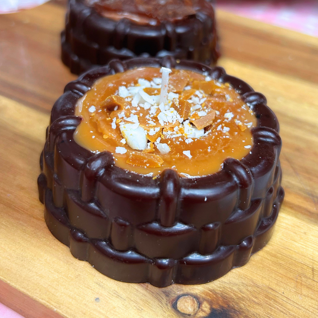 A close-up of a round chocolate candle filled with caramel-colored caramel or custard, topped with white chocolate shavings and a small wick, sitting on a wooden board with another similar mold blurred in the background.