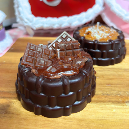 Two novelty chocolate-themed candles shaped like chocolate cake layers, one with dark chocolate frosting and chocolate bar decorations, and another in the background with caramel frosting and shredded coconut, displayed on a wooden surface.