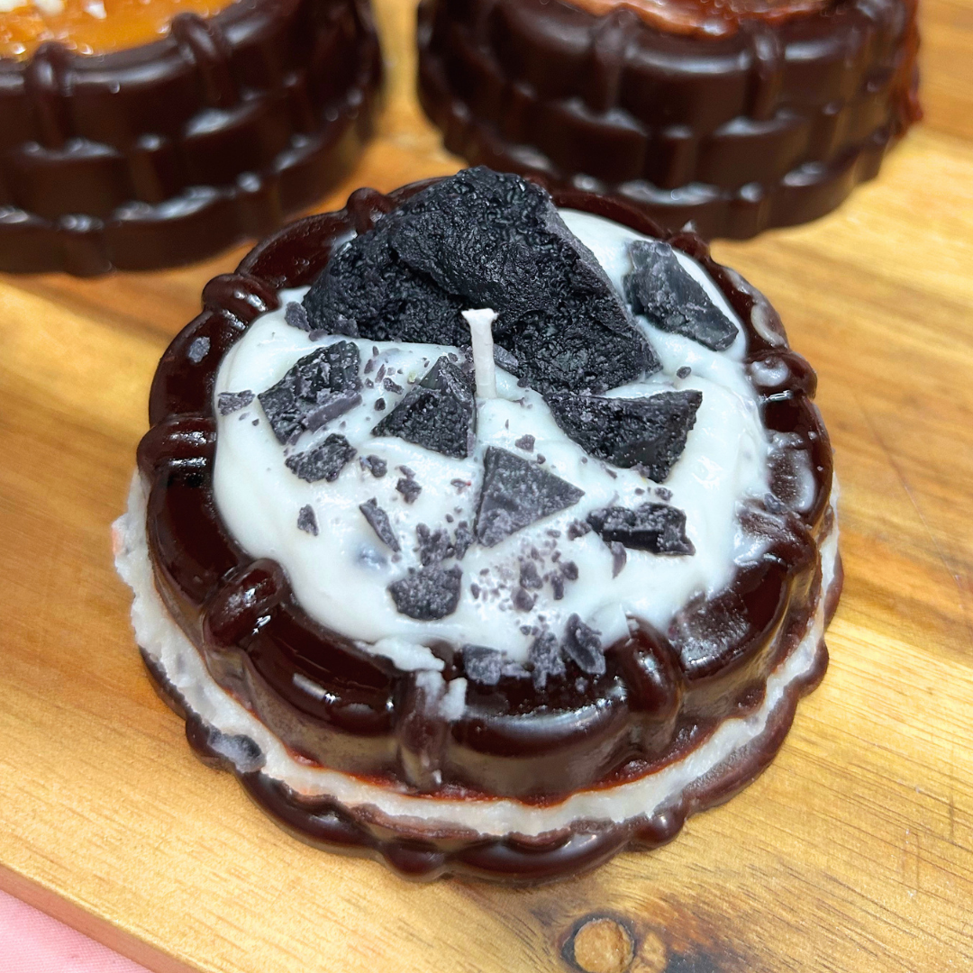 A round novelty candle styled like a chocolate cake, with dark brown chocolate layers, a white cream center, and crushed dark cookie pieces on top, sitting on a wooden surface.