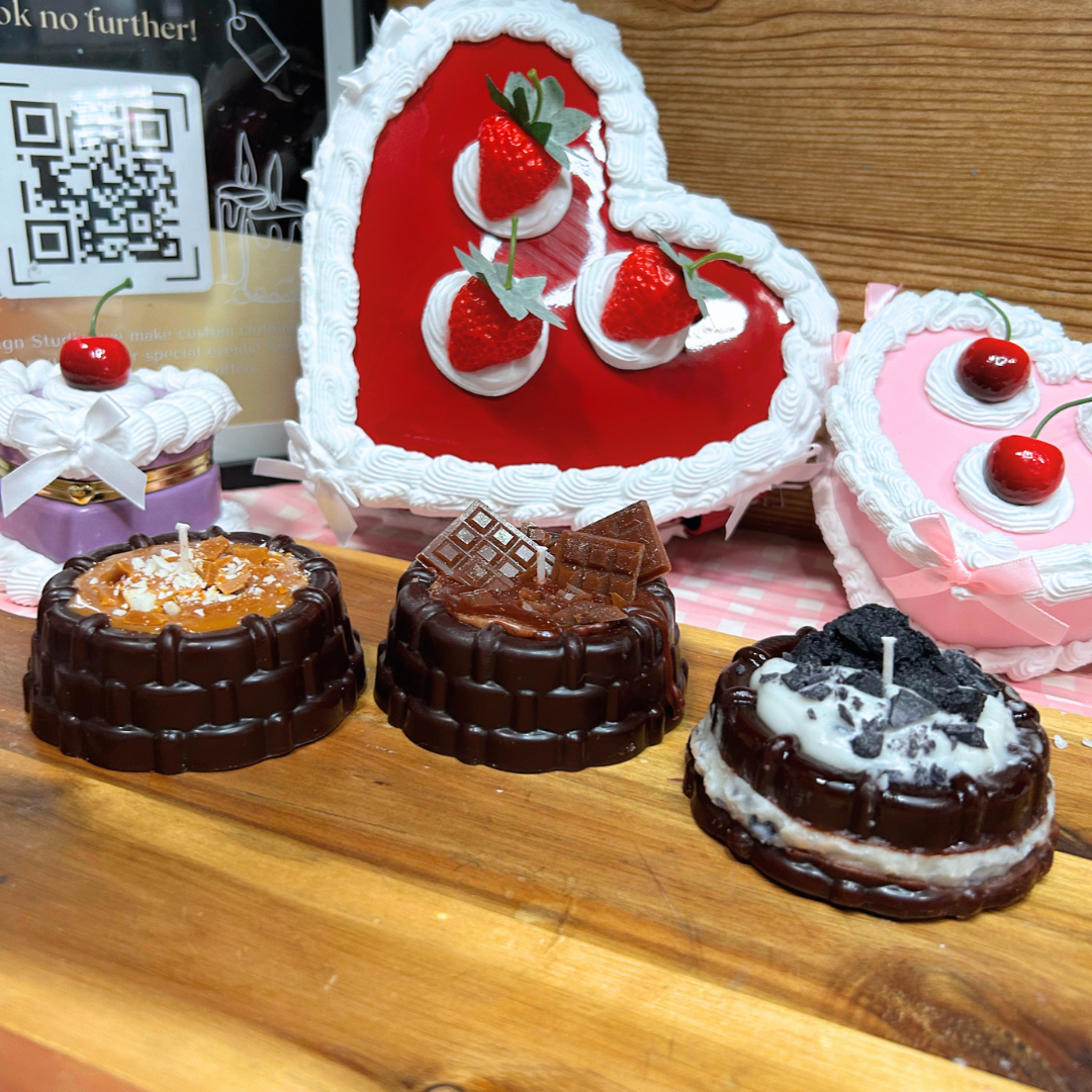 A set of heart-shaped and round candles styled like desserts, including chocolate cake layers, strawberry cream pie, and pink cake with cherries, arranged on a wooden board.