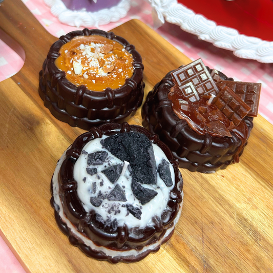 Three miniature brownie shortcakes arranged on a wooden board. One cup is topped with caramel-colored chocolate and white shavings, while the other two are dark chocolate with white and black chocolate toppings.