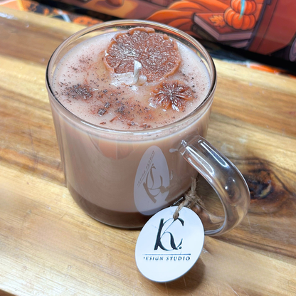 A clear glass mug candle filled with light brown wax, topped with a dried citrus slice and dark speckled wax, sitting on a wooden surface. A white oval tag with a black logo and the text 'DESIGN STUDIO' hangs from the handle.