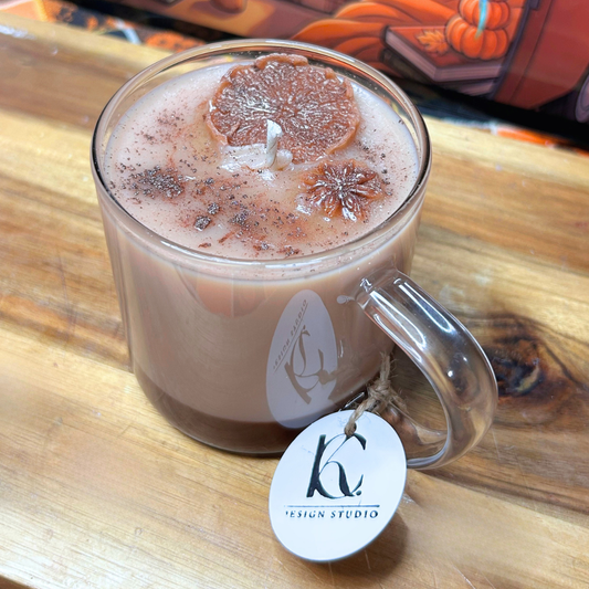 A clear glass mug candle filled with light brown wax, topped with a dried citrus slice and dark speckled wax, sitting on a wooden surface. A white oval tag with a black logo and the text 'DESIGN STUDIO' hangs from the handle.