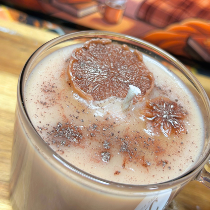 A close-up of a chai latte candle in a clear glass mug, topped with a dried orange slice, a star anise, and a dusting of cinnamon powder, set on a wooden surface.