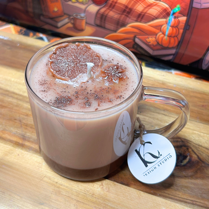 A clear glass mug chai candle filled with a brown design topped with a dried citrus slice and cocoa powder, sitting on a wooden surface. A round white tag with a black 'KC' logo hangs from the handle, and a colorful illustrated background is visible behind it.