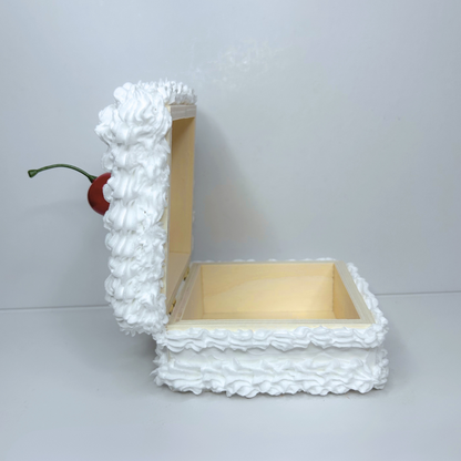Side view of open square wooden storage box with frosted cake design and cherry on top.
