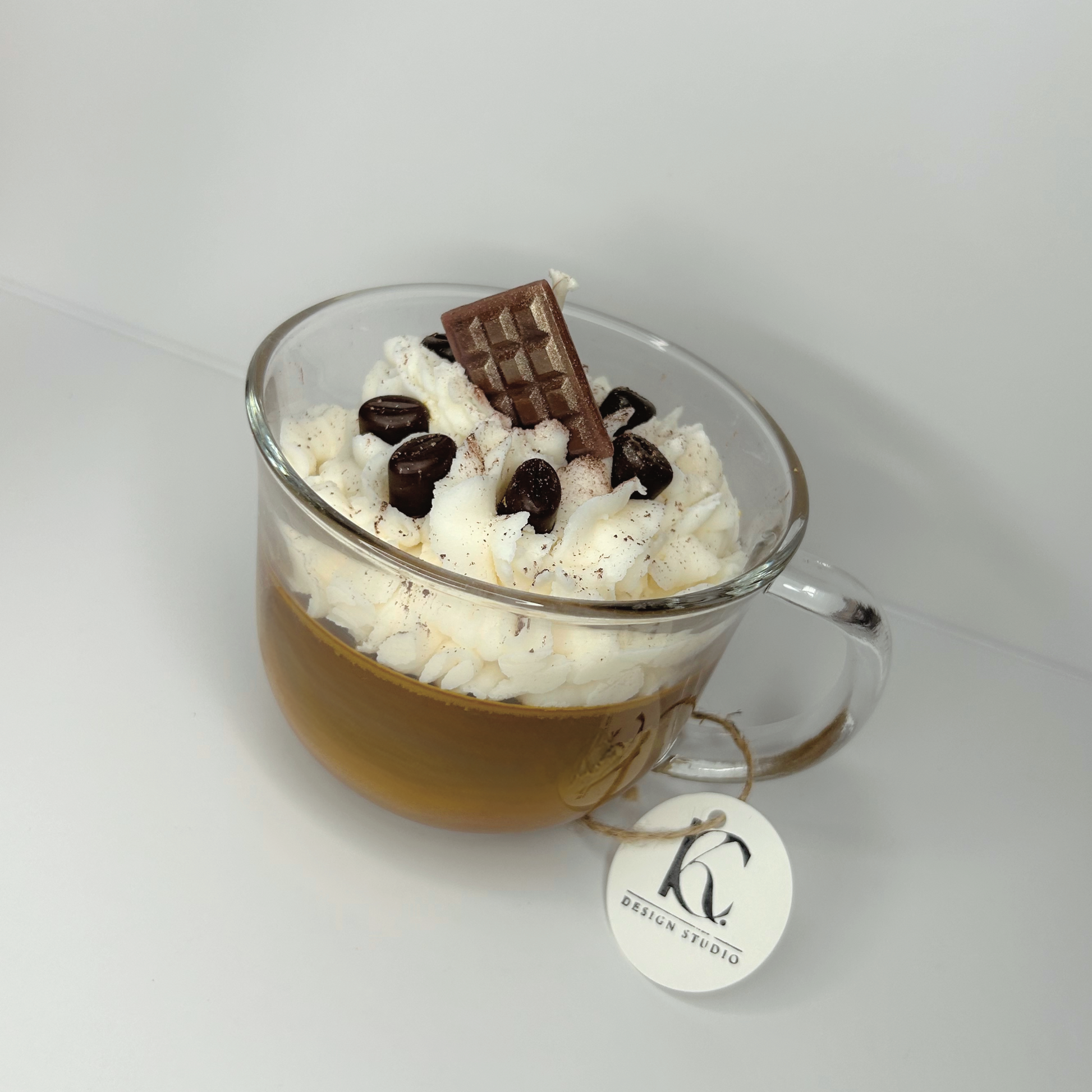 Angled view of Chocolate Vanilla Latte Candle in an 18oz glass mug, showing the layers of whipped cream, mini coffee beans, and a chocolate bar on top, with a hint of the rich coffee house, vanilla buttercream, and salted caramel scents.