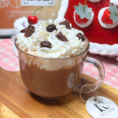 A clear glass mug candle made with a coffee-style design topped with whipped cream, chocolate pieces, and a small candle, set on a wooden surface with a strawberry-themed background.
