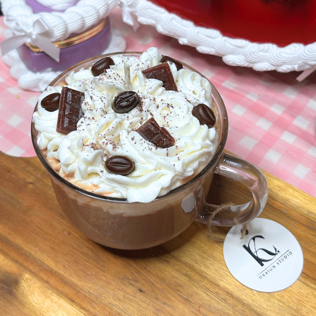 A clear glass mug candle made with a coffee-inspired drink design topped with whipped cream, chocolate chunks, and coffee beans, resting on a wooden surface with a pink-and-white check cloth and a round 'KC Design Studio' tag.