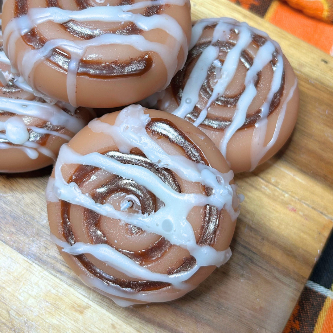 Four cinnamon roll candles with white icing drizzle on a wooden board.