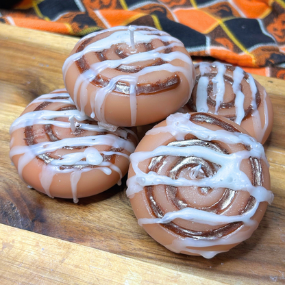 Four cinnamon roll-shaped candles arranged on a wooden board, with white drizzle details and a patterned orange, black, and cream cloth in the background.