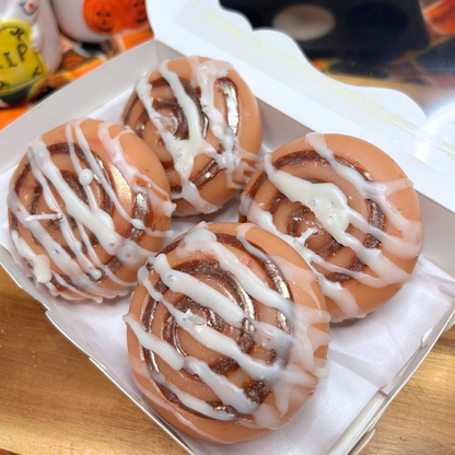 Four cinnamon roll candles with white icing drizzle in a white cardboard box, photographed close-up on a wooden surface.