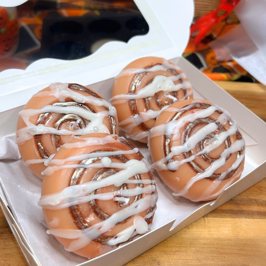 Four cinnamon roll candles with white icing drizzle in a white cardboard box on a wooden surface.