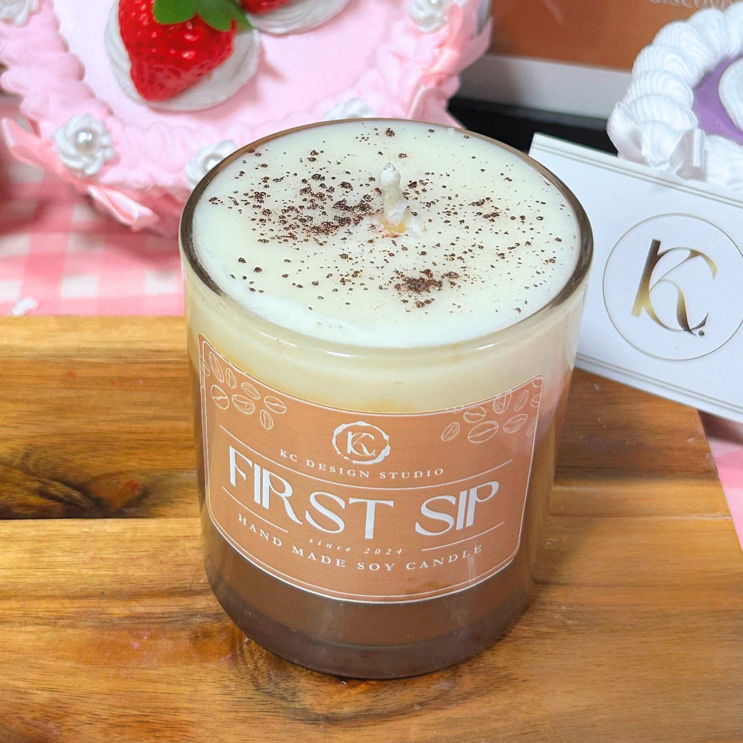 A glass jar candle labeled 'FIRST SIP' by KC Design Studio sits on a wooden surface. The candle has a creamy off-white wax top with dark brown speckles and a warm peach-brown label.