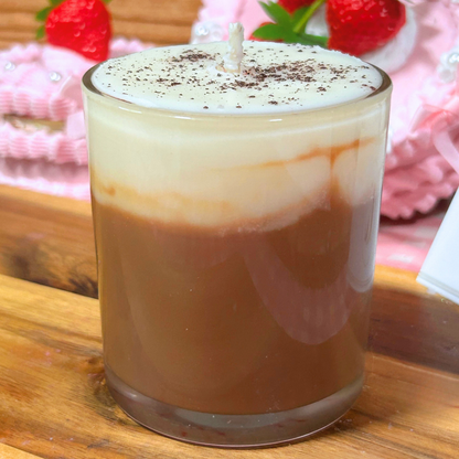 A glass jar candle styled like a layered coffee drink, with a creamy white top layer dusted with cocoa powder, set on a wooden surface with strawberries in the background.