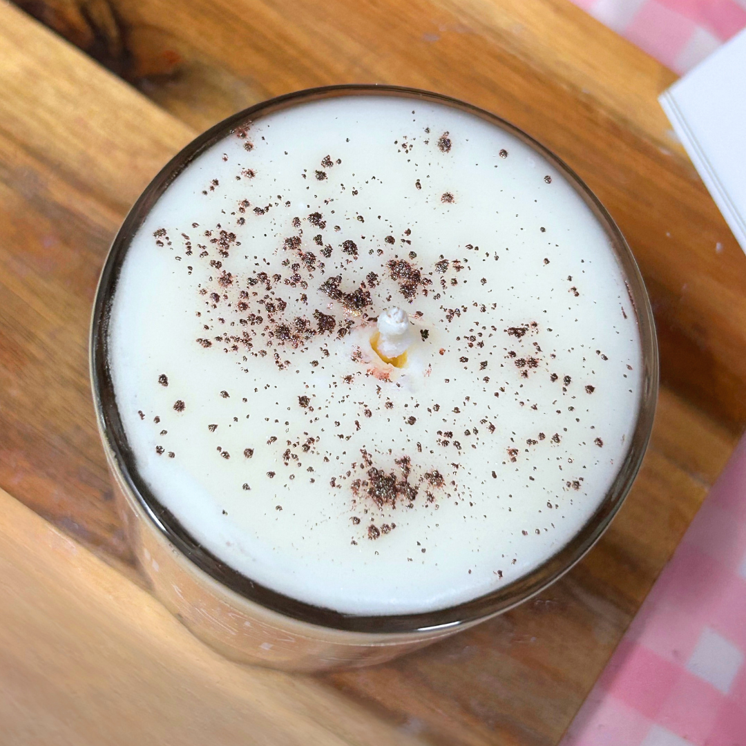 Top-down view of a white candle in a clear glass jar, topped with dark brown cocoa powder, resting on a wooden board.