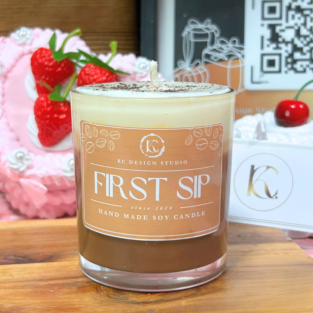A soy candle in a clear glass jar with a frosted top and a tan label reading 'FIRST SIP' and 'KC DESIGN STUDIO'. The candle sits on a wooden surface with decorative strawberries and gift boxes in the background.
