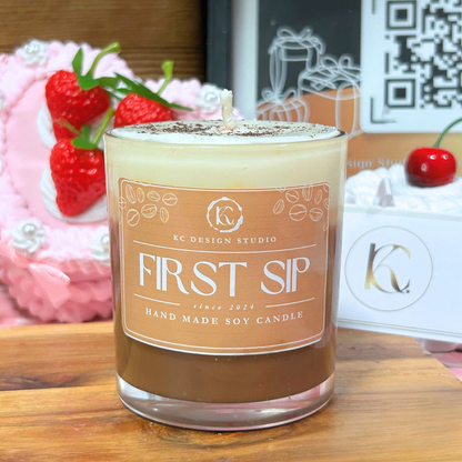 A soy candle in a clear glass jar with a frosted top and a tan label reading 'FIRST SIP' and 'KC DESIGN STUDIO'. The candle sits on a wooden surface with decorative strawberries and gift boxes in the background.