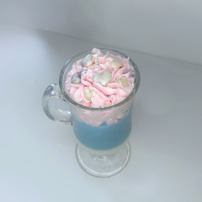 Angled view of cotton candy candle with blue and white base, pink topping, and decorative sprinkles in a clear glass mug.