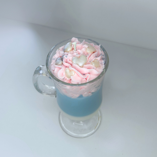 Angled view of cotton candy candle with blue and white base, pink topping, and decorative sprinkles in a clear glass mug.
